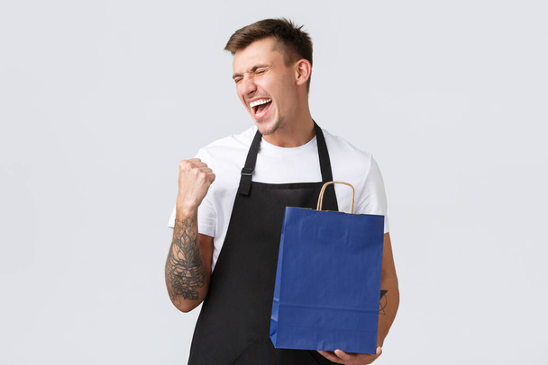 Retail store, shopping and employees concept. Cheerful happy salesman enjoying finally working after covid-19, fist pump and singing from happiness, holding eco-bag, white background - Photo, Image