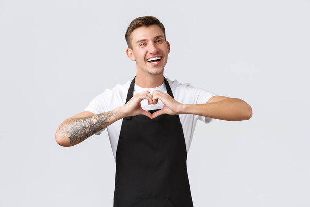 Employees, grocery stores and coffee shop concept. Friendly happy waiter in black apron loving visitors, inviting visit cafe and enjoy drinks, showing heart love sign, white background - Photo, Image