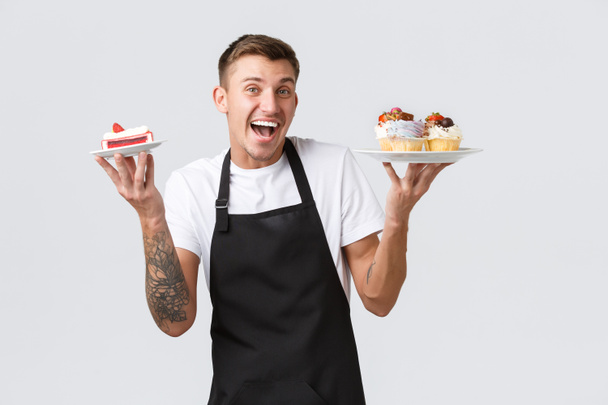 Mały handel detaliczny, kawiarnie i koncepcja piekarni. Entuzjastycznie przystojny barista, kelner trzymający babeczki i ciasto, sprzedający desery klientom, uśmiechający się optymistycznie na białym tle - Zdjęcie, obraz
