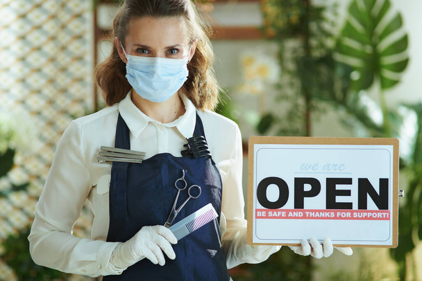 Small business survival after covid-19 pandemic. Portrait of elegant small business owner woman in apron with medical mask, gloves, hair comb and scissors showing open sign. - Foto, Bild