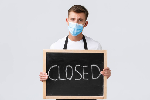 Small retail business owners, covid-19 and social distancing concept. Gloomy and upset salesman, waiter in medical mask inform that cafe closed, showing sign and frowning disappointed - Foto, imagen