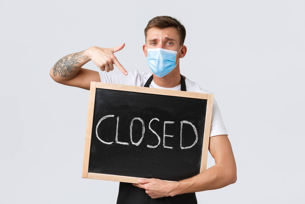 Small retail business owners, covid-19 and social distancing concept. Disappointed and sad salesman in medical mask, barista showing closed sign, inform clients restaurant not working - Foto, Bild