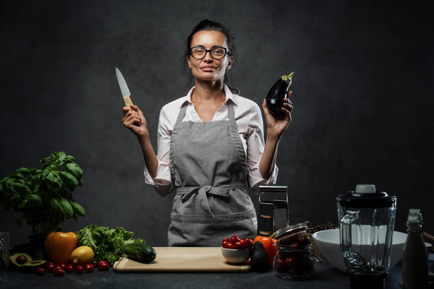 Piękne dojrzałe kobieta gotowanie w kuchni, pozowanie gospodarstwa nóż i bakłażan - Zdjęcie, obraz
