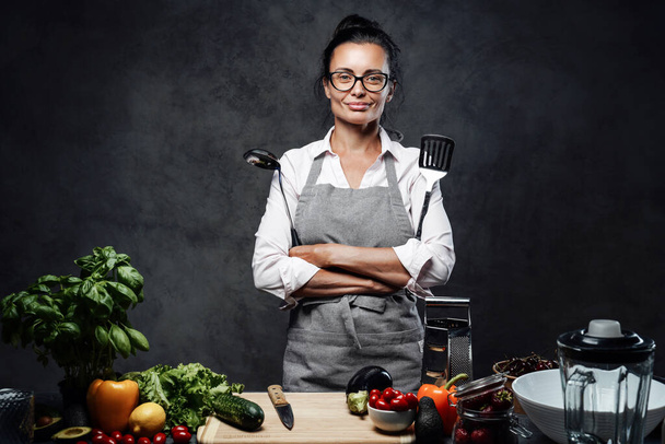 Bella cuoca di mezza età in posa accanto a un tavolo con verdure fresche in cucina - Foto, immagini