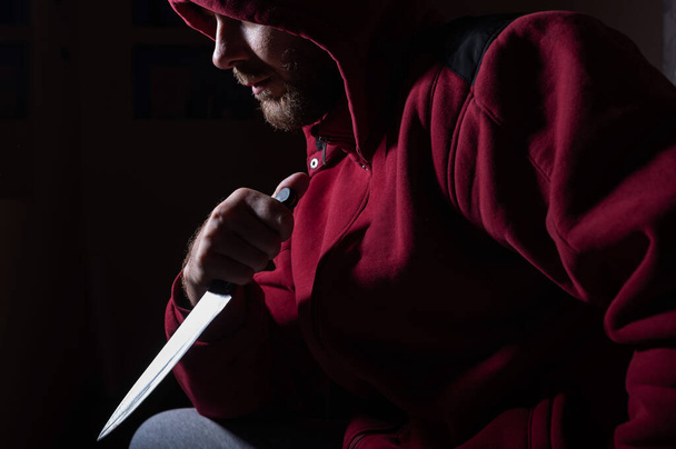 Опасный бородатый мужчина в красном капюшоне держит острый нож у лица. Преступник или убийца с ножами в темноте. - Фото, изображение