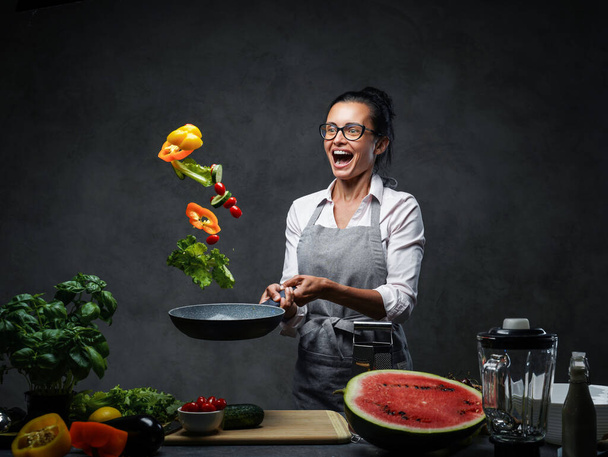Emocional mujer madura chef lanzando verduras picadas de una sartén
 - Foto, Imagen