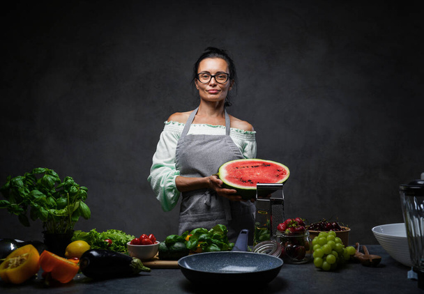 Piękna dojrzała kobieta gotowanie w kuchni, posiada połowę arbuza i patrząc na aparat - Zdjęcie, obraz