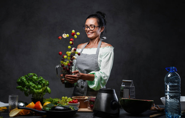 Felice chef di mezza età che lancia frutta fresca nell'aria da un frullatore in cucina - Foto, immagini