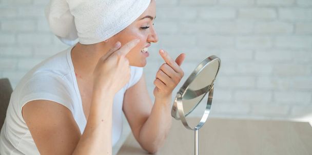 An upset woman looks in the mirror and squeezes a pimple on her cheek. A girl with hormonal problems does facial cleansing at home. Teen acne. Himself a dermatologist in quarantine. Problem skin. - Фото, изображение