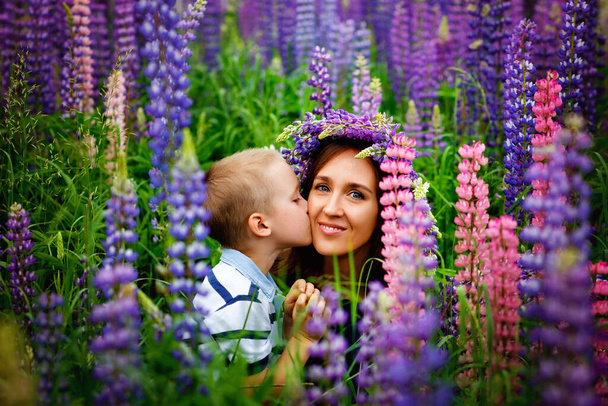 Een moeder in een blauwe jurk met een krans van bloemen op haar hoofd houdt haar zoon in haar armen in een bloeiend veld van lila Lupin bloemen. Zoon kust moeder op de wang - Foto, afbeelding