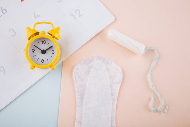 Calendrier, tampon de coton et réveil jaune sur fond bleu rose. Concept de cycle menstruel féminin - Photo, image
