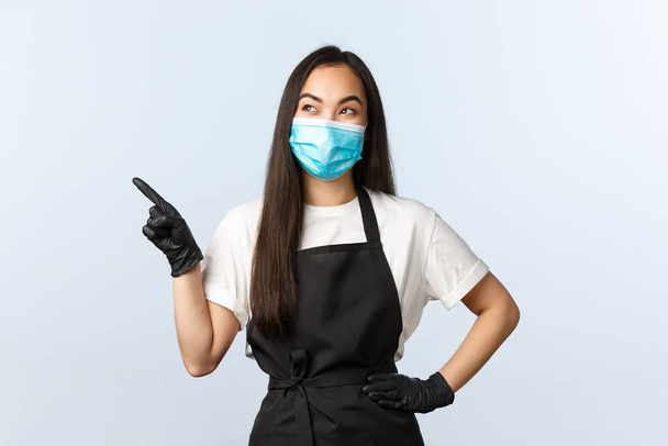 Covid-19 πανδημία, κοινωνική αποστασιοποίηση, μικρές επιχειρήσεις και πρόληψη της έννοιας του ιού. Χαρούμενη χαμογελαστή Ασιάτισσα μπαρίστα καφετέριας με ιατρική μάσκα και γάντια βλέποντας καλό promo στην πάνω αριστερή γωνία - Φωτογραφία, εικόνα