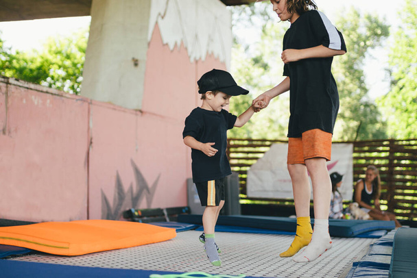 Grote broer leert jongere om op een trampoline te springen. Kinderen actief vrijetijdsbesteding trampolining hand in hand. Sportweekend in het sportcentrum van acrobatiek en trampoline. Lichamelijk onderwijs. - Foto, afbeelding