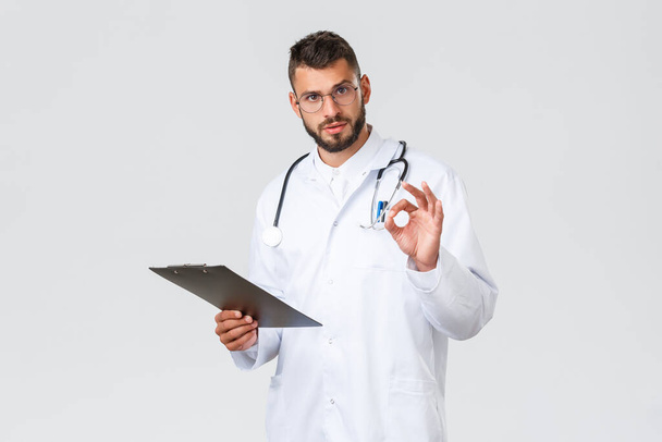 Healthcare workers, medical insurance, clinic lab and covid-19 concept. Handsome serious doctor in white coat, glasses and clipboard, show okay sign, assure tests alright, results are good - Foto, Imagen