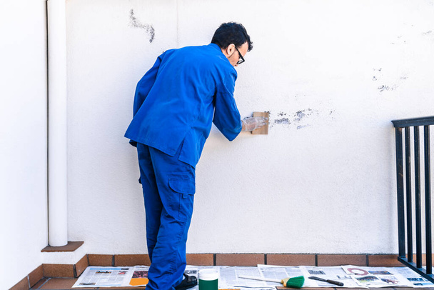 Schilder schrobben de witte muur van het huis met schuurpapier. Volwassen man schilderen en hervormen van het terras van het huis. - Foto, afbeelding
