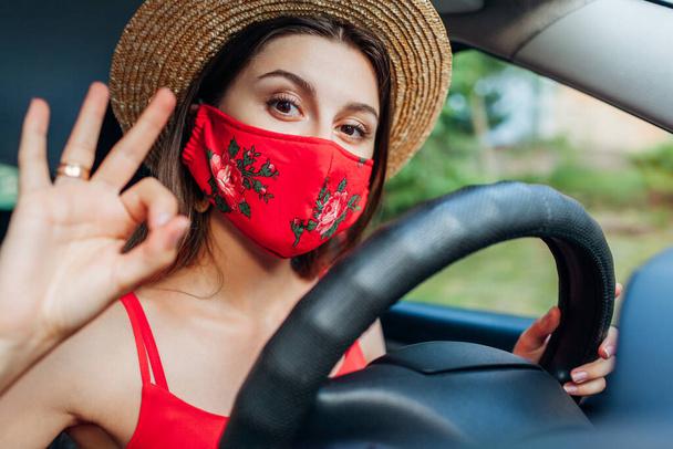 Conductora sentada en coche con máscara protectora durante la pandemia de coronavirus. Chica con estilo en la ropa de verano, accesorios muestra ok signo fresco
 - Foto, Imagen