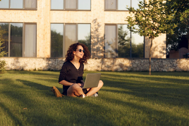 Πορτρέτο της όμορφης νεαρής γυναίκας με γυαλιά ηλίου κάθεται στο πράσινο γρασίδι με τα πόδια σταυρωμένα κατά τη χρήση laptop χαμογελώντας. - Φωτογραφία, εικόνα