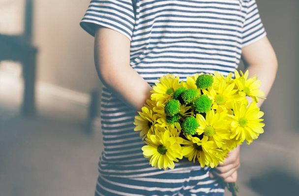 Rear view of kid with bunch of beautiful yellow Chrysanthemum behind his back preparing nice surprise for his mother or Father, Child in pajamas holds flowers behind his back, Romantic or Mother day - Photo, Image