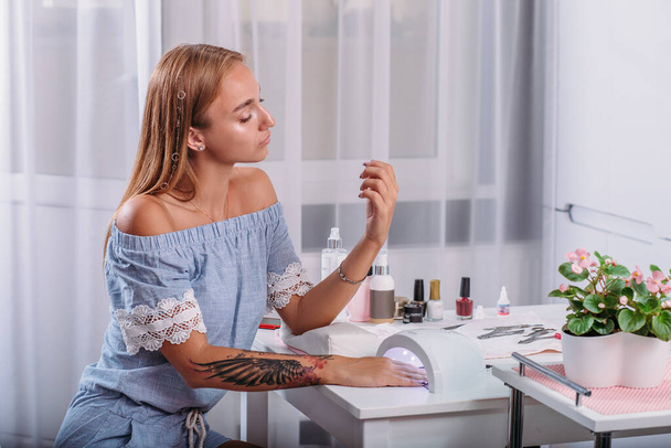 Beautiful girl with tattoo sits at the table and dries under the LED lamp fresh manicure and nail varnish - Foto, afbeelding