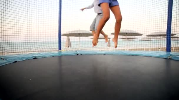 Lähellä jalat kaksi lasta hyppää trampoliini rannalla auringonlaskun aikana. Lapsuuden ja aktiivisen perheelämäntavan käsite. Poika ja tyttö hauskaa kesälomaa merellä. - Materiaali, video