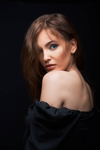 portrait of a beautiful woman in a dark dress posing on a dark background, beautiful brown hair, blue eyes looking from shoulder - Photo, Image