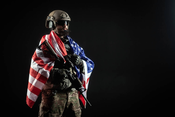 銃と軍事制服を着たアメリカの兵士は、暗い背景、アメリカのエリート部隊、アメリカの特殊部隊に対して米国の旗を保持しています。 - 写真・画像