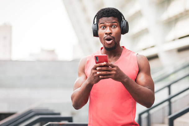 verrast Afrikaans-Amerikaanse man luisteren naar muziek op koptelefoon tegen de achtergrond van de stad in de zomer, geschokt man met een smartphone, kopieer ruimte - Foto, afbeelding