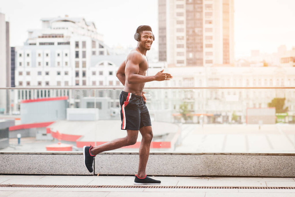 Afro-Amerikaanse sportman rent snel vooruit en luistert naar muziek tegen de achtergrond van de stad in de zomer, atletische man runner trein en glimlach - Foto, afbeelding