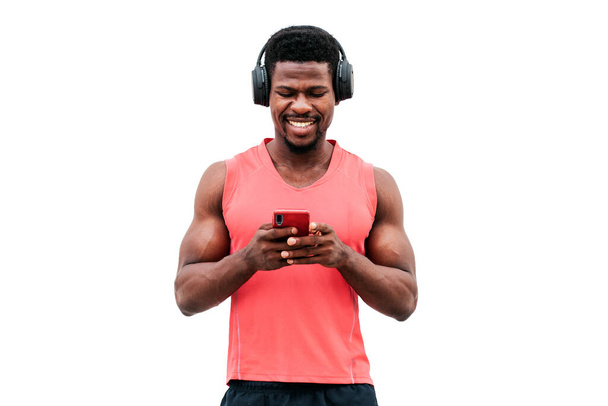 african american athletic guy holds a smartphone and listens to music on a white isolated background, a man with headphones is looking at the phone and smiling - Photo, Image