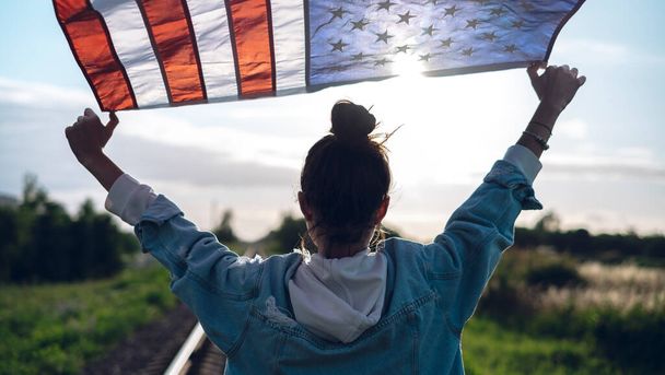 Portrait of a young beautiful woman in glasses and jeans with a waving American flag in her hands, background of blue sky, concept of patriotism, demonstration, protest. - Фото, изображение