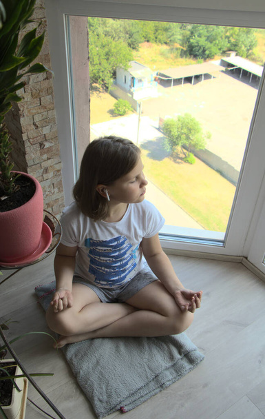 Fülhallgatós lány, lótuszpózban ül az ablaknál. A gyerek otthon jógázik. A kilátás az ablakból az úton. Egy gyerek ül a padlón, felülnézetből. - Fotó, kép