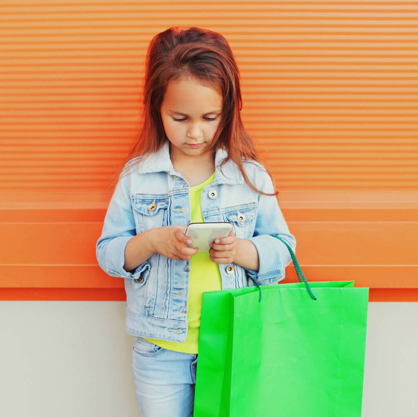 little girl child with phone and shopping bags on city street over orange wall background - Photo, image