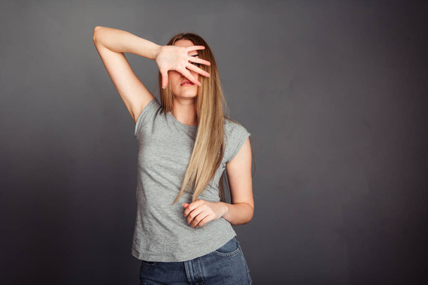 Chica de pelo rubio cubre su cara con los dedos separados en una camiseta de fondo gris liso y jeans
 - Foto, imagen