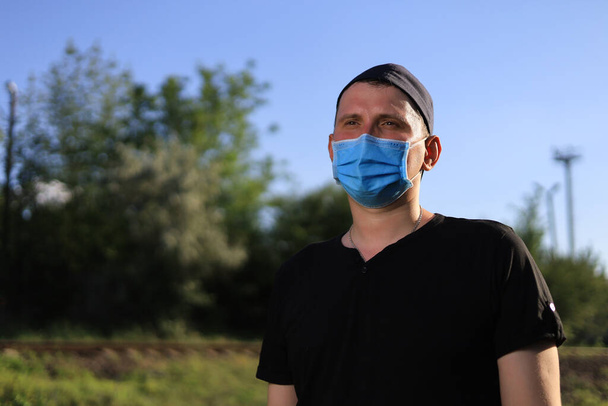 Caucasian young man or adult guy in casual clothes and a medical surgical mask is resting in a city park. - Photo, Image