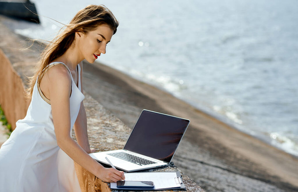 όμορφη, σοβαρή γυναίκα σε ένα λευκό φόρεμα με ένα δισκίο, στυλό, φορητό υπολογιστή τηλέφωνο στην αποβάθρα - Φωτογραφία, εικόνα
