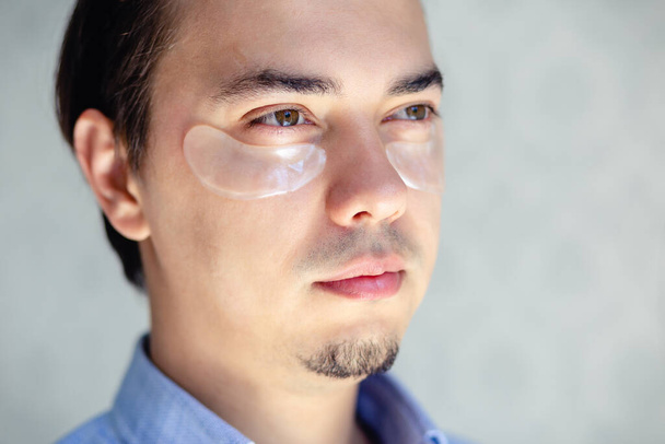 Close up Handsome man looking aside with eye patch preventing from dark circles, taking care of his face skin on light background. Men's personal care.  - Photo, Image
