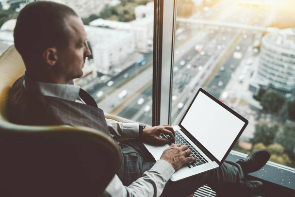 Side view of a stately successful man entrepreneur with a laptop with white blank screen mock-up, sitting on an armchair on the top floor of a luxury business office skyscraper with a highway outside - Fotó, kép