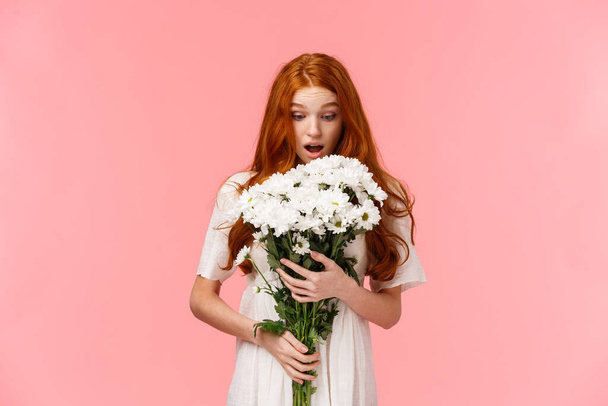 Surprised attractive redhead girl receive romantic valentines day gift, looking at beautiful bouquet of flowers amazed and wondered who might sender be, standing over pink background - Fotó, kép