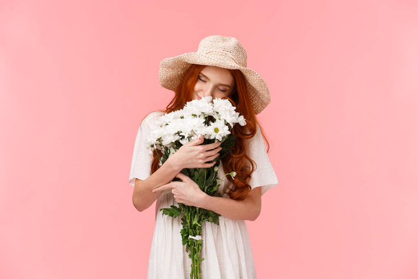 Romance, love and relationship concept. Romantic dreamy, alluring redhead girl sniffing flowers, close eyes and hugging beautiful bouquet, standing pink background - Fotoğraf, Görsel