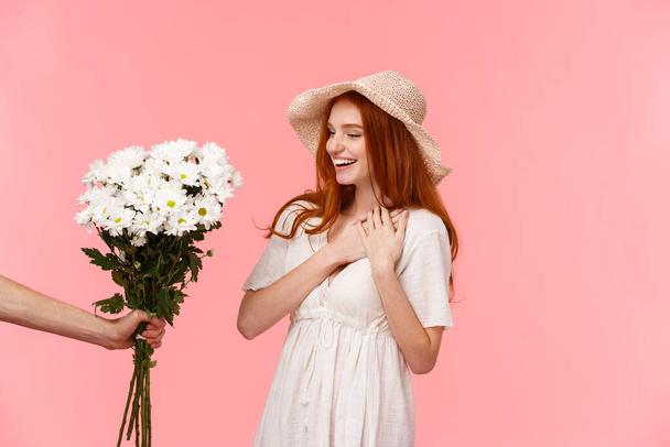 Gifts, celebration and tenderness concept. Surprised cute, alluring redhead woman in hat, dress, charmed with pleasant gift, holding hands on heart flattered, smiling, looking at bouquet flowers - Photo, Image