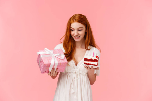 Curieux et heureux, joyeux attrayant rousse fille en robe blanche, célébrer l'anniversaire, recevoir un cadeau mignon, en regardant cadeau, en gardant la paix du gâteau avec une bougie allumée, debout fond rose - Photo, image