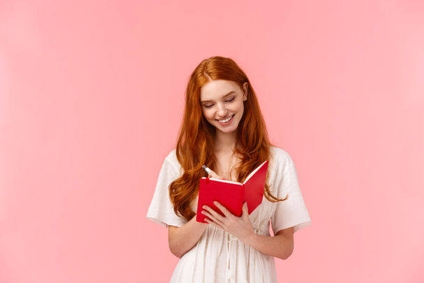Lovely, sensual and stylish redhead girl making list, studying, writing homework or schedule in cute red notebook. smiling dreamy looking at notes, standing pink background delighted - Photo, Image