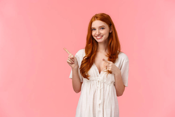 Waist-up portrait lovely european redhead female in white fashionable dress, pointing finger upper left corner, invite come and check-out promotion, give recommendation, pink background - Foto, Imagem