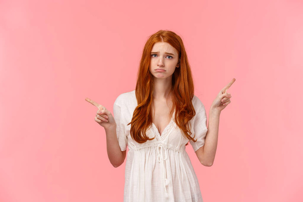 Upset, pouting and gloomy silly redhead female in white dress, whining, telling about bad experience, pointing fingers upper right and left corners, two variants both didnt fit, pink background - Photo, Image