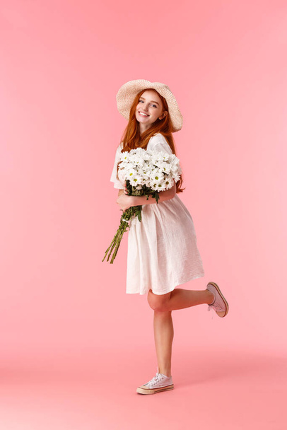 Full-length κάθετη βολή ευτυχισμένη, χαρούμενη ελκυστική γυναίκα με κόκκινα μακριά μαλλιά, ψάθινο καπέλο, φορώντας χαριτωμένο καλοκαιρινό λευκό φόρεμα, χορός, πηγαίνοντας σπίτι από ρομαντική ημερομηνία με λουλούδια, κρατώντας μπουκέτο - Φωτογραφία, εικόνα