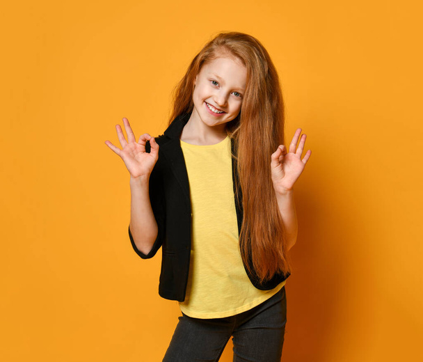Ginger teenage girl in black jacket, pants, yellow t-shirt. She smiling, showing okay sign, posing on orange background. Close up - Foto, Imagem