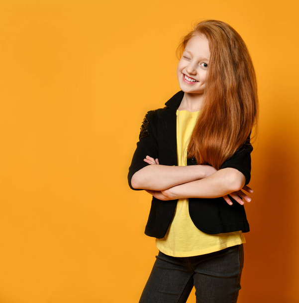 Red-haired teenage girl in black jacket, pants, yellow t-shirt. She smiling, hands folded, posing on orange background. Close up - Foto, Imagem