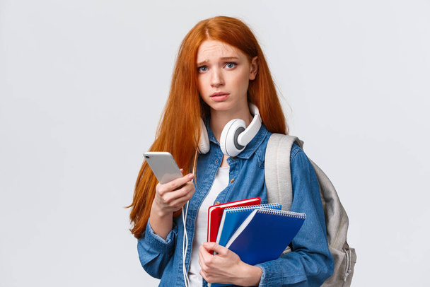 Uneasy, sconvolto e cupo carino rossa adolescente ragazza, studente in università accigliato sguardo angosciato e infelice fotocamera, tenendo zaino educazione notebook e smartphone - Foto, immagini