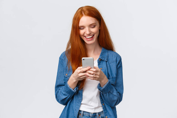 Chatten, communicatie en mobiel concept. Vrolijke roodharige glimlachende meisje sms 'en vriend, lachen als het kijken naar grappige vlog, op zoek smartphone display blij, staande witte achtergrond - Foto, afbeelding
