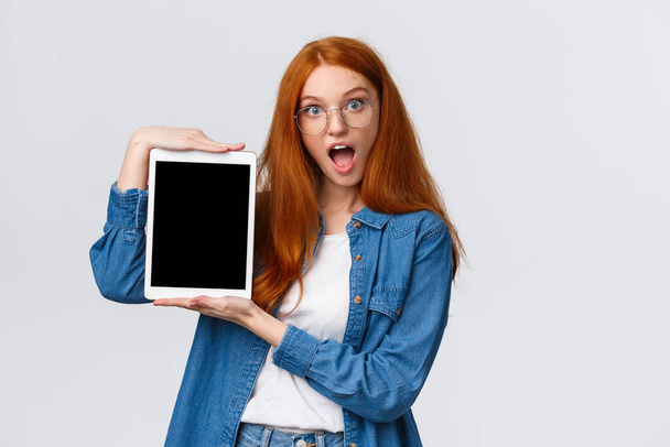 Excited and amused attractive redhead teenage girl making design, creative project on device screen, showing digital tablet display, look amazed, open mouth fascinated, white background - Foto, afbeelding